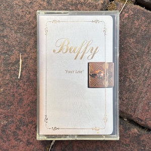 May include: A vintage cassette tape with a white cover featuring the gold-colored word "Buffy" and the text "FIRST LOVE". The tape has a decorative gold border and a small, ornate clasp.