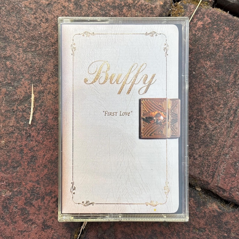 May include: A vintage cassette tape with a white cover featuring the gold-colored word "Buffy" and the text "FIRST LOVE". The tape has a decorative gold border and a small, ornate clasp.