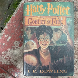 Puede incluir: Un libro de tapa dura verde, naranja y dorado con el título "Harry Potter y el Cáliz de Fuego" y el autor "J.K. Rowling". La portada presenta una ilustración de Harry Potter con una varita mágica, rodeado de otros personajes.