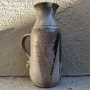 May include: A tall, white ceramic vase with a brown glaze and a handle. The vase has a textured surface and a slightly flared opening.