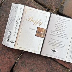 May include: A cassette tape and its case are open, revealing the album art for "Buffy First Love." The case is white with gold lettering and a decorative clasp. The cassette tape is labeled "VEL 0214."