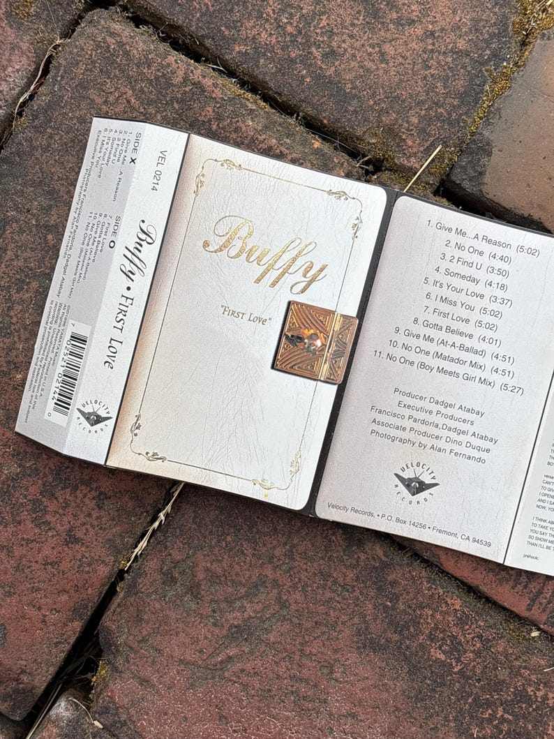 May include: A cassette tape and its case are open, revealing the album art for "Buffy First Love." The case is white with gold lettering and a decorative clasp. The cassette tape is labeled "VEL 0214."