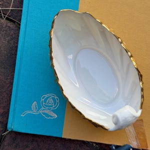 May include: An oval-shaped white porcelain dish with a scalloped edge and a metallic gold rim. The dish features a smooth, glossy interior surface and a curved handle at one end, showcasing a refined design with clean lines and a polished finish.