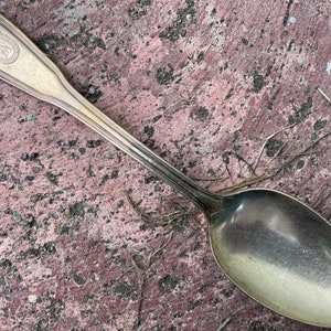 Peut inclure: Une cuillère en argent avec un manche orné et un monogramme sur le dessus. La cuillère est posée sur une surface rose rugueuse.