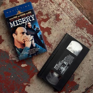May include: A VHS tape and its cover for the movie "Misery" are displayed on a weathered surface. The cover features James Caan and Kathy Bates, with the title in large white letters. The VHS tape is black with white labels.