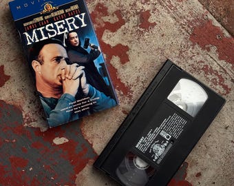 Może przedstawiać: Kaseta VHS i jej okładka do filmu "Misery" są wyświetlane na zniszczonej powierzchni. Okładka przedstawia Jamesa Caana i Kathy Bates, z tytułem dużymi białymi literami. Kaseta VHS jest czarna z białymi etykietami.