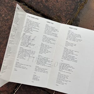 May include: A white paper insert with song lyrics, including "It's Your Love," "I Miss You," and "First Love." The lyrics are printed in black text. The paper is folded and lies on a textured, dark surface.
