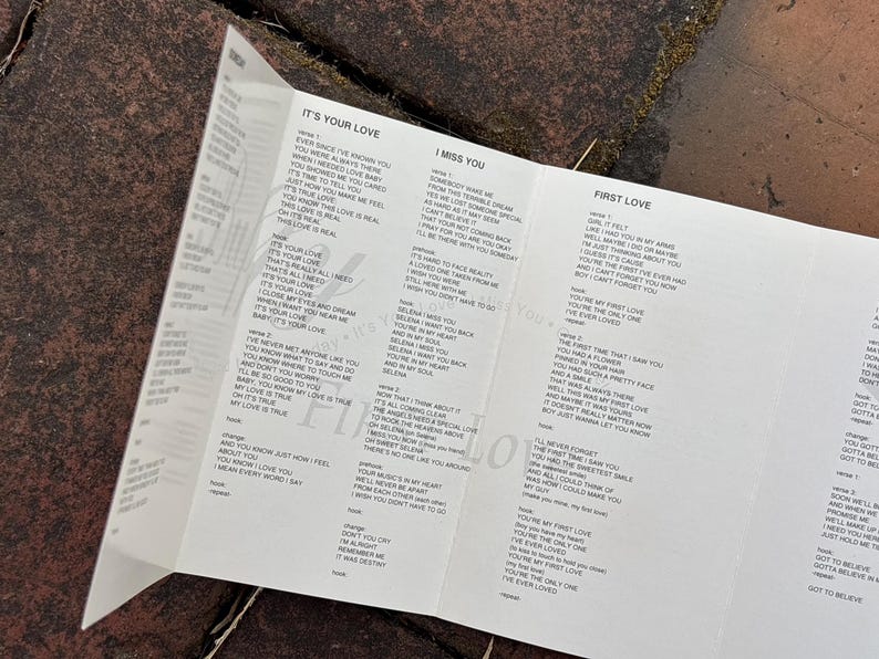 May include: A white paper insert with song lyrics, including "It's Your Love," "I Miss You," and "First Love." The lyrics are printed in black text. The paper is folded and lies on a textured, dark surface.