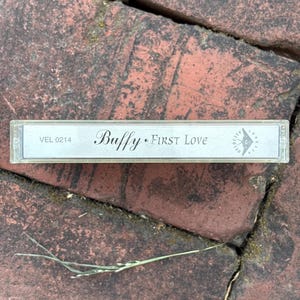 May include: A vintage cassette tape labeled "Buffy First Love" in a clear plastic case. The tape is set against a background of red bricks. The text "VEL 0214" is also visible on the tape.