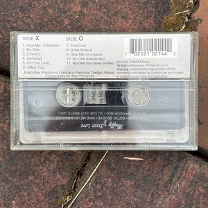 May include: A clear cassette tape with the album title "First Love" by Buffy. The cassette case has track listings for Side X and Side O. The tape is resting on a textured, reddish-brown surface.