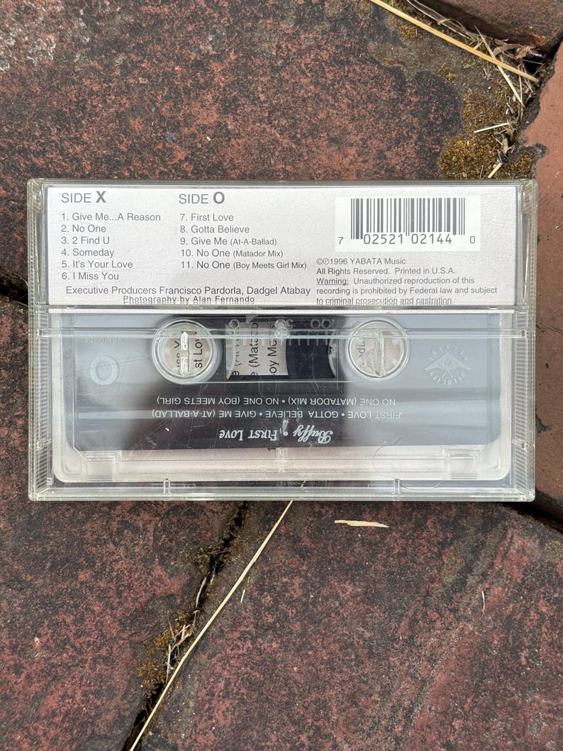 May include: A clear cassette tape with the album title "First Love" by Buffy. The cassette case has track listings for Side X and Side O. The tape is resting on a textured, reddish-brown surface.