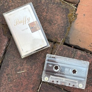 May include: A clear cassette tape and case with the title "Buffy" and "First Love" printed on the case. The cassette tape is visible, with its internal components and labels. The case has a gold-colored design.
