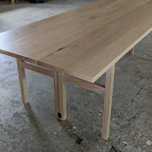 May include: A light-colored wooden table with a rectangular top and four legs. The table appears to be made of solid wood, with visible wood grain. The table is approximately 60 inches long and 30 inches wide.