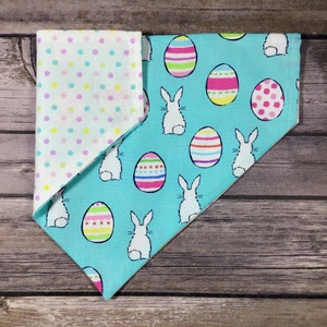 May include: Reversible pet bandana with an Easter theme. One side features a light blue background with white bunnies and colorful Easter eggs. The other side has a white background with pastel polka dots. The bandana is made of fabric.