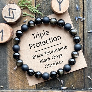 May include: A black beaded bracelet with the text "Triple Protection" and the words "Black Tourmaline, Black Onyx, Obsidian" on a tan card. The bracelet is made of black beads, some with a golden sheen. The background is a wooden surface.