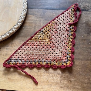 May include: A handmade crochet bandana in a triangular shape. The bandana features a gradient of colors, including shades of pink, yellow, and brown. The edges are finished with a scalloped border in a deep red color, and it has ties for securing.