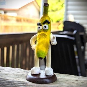 May include: A whimsical, hand-painted banana figurine with a grumpy expression. The yellow banana has a green tip and is wearing white shoes and gloves. It stands on a brown circular base, with one arm pointing. The figurine is a unique decorative item.