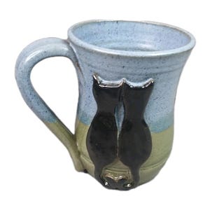 May include: A handmade ceramic mug with a light blue and green glaze. Two black cat silhouettes are attached to the side of the mug. The mug has a curved handle and a wide opening.