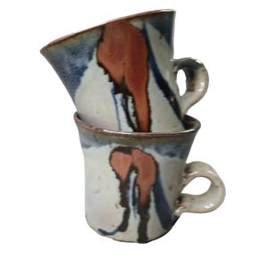 May include: Two stacked ceramic mugs with abstract designs. The mugs have a cream-colored base with brown, blue, and orange brushstrokes. Each mug has a curved handle and a slightly flared rim.