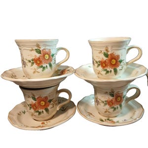 May include: Four cream-colored teacups and saucers. Each cup and saucer features a floral design with orange and purple flowers and green leaves. The cups have curved handles and sit atop matching saucers.