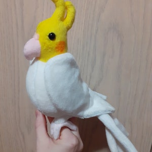 Puede incluir: Un juguete de peluche de cacatúa blanco y amarillo con un pico rosa y ojos negros. El pájaro está sentado en una mano.