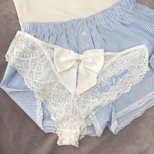 May include: White lace underwear featuring a large white bow and the embroidered text "Mrs. Orona". The lingerie is paired with blue and white striped pajama shorts and a white top. The design is delicate and feminine.
