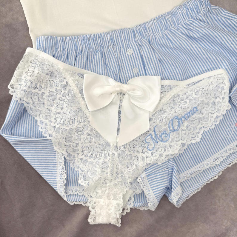 May include: White lace underwear featuring a large white bow and the embroidered text "Mrs. Orona". The lingerie is paired with blue and white striped pajama shorts and a white top. The design is delicate and feminine.