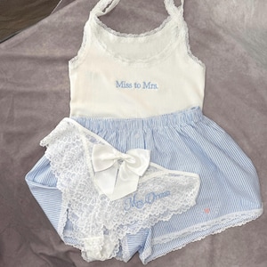 May include: A white vest top with the embroidered text "Miss to Mrs." is paired with blue and white striped shorts and a matching lace-trimmed underwear set. A white bow adorns the underwear.