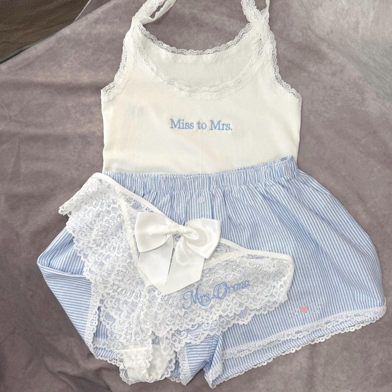 May include: A white vest top with the embroidered text "Miss to Mrs." is paired with blue and white striped shorts and a matching lace-trimmed underwear set. A white bow adorns the underwear.