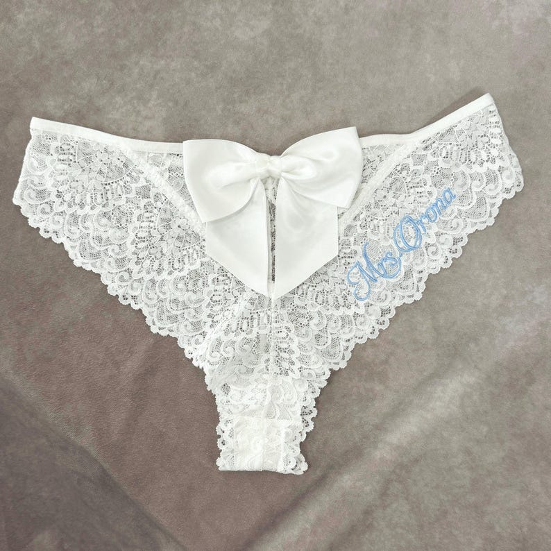 May include: White lace underwear featuring a large satin bow. The underwear has a delicate floral lace pattern and the words "Mrs. Orona" are embroidered in light blue.