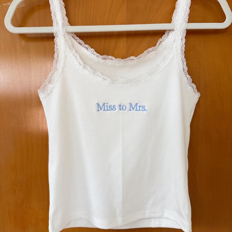 May include: A white camisole top with lace detailing on the straps and neckline. The words "Miss to Mrs." are embroidered in light blue. The top is displayed on a white hanger.