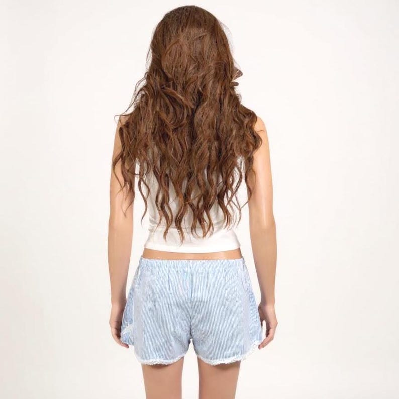 May include: A white sleeveless top paired with light blue and white pinstriped shorts featuring a delicate white lace trim along the bottom hem. The outfit is displayed on a mannequin with long, wavy, light brown hair viewed from behind.