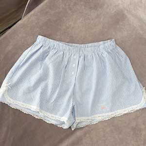 May include: Pale blue and white striped shorts with a button closure and elasticated waistband. The shorts feature white lace trim and a small pink embroidered design near the hem.