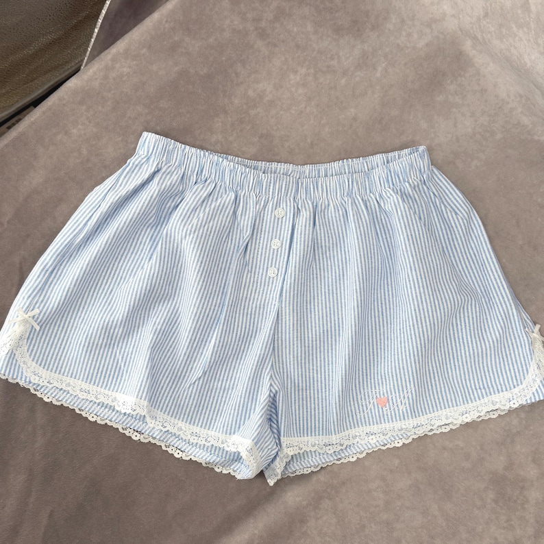 May include: Pale blue and white striped shorts with a button closure and elasticated waistband. The shorts feature white lace trim and a small pink embroidered design near the hem.
