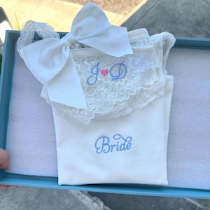 May include: A white bridal set, including a top with lace trim and a large bow, presented in a blue box. The top is embroidered with "Bride" in blue script and initials "J & D" with a pink heart.