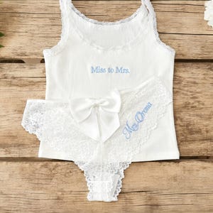 May include: White bridal lingerie set. A tank top with "Miss to Mrs." in light blue, and lace trimmed panties with a bow and "Mrs. Orona". The set is displayed on a wooden surface.