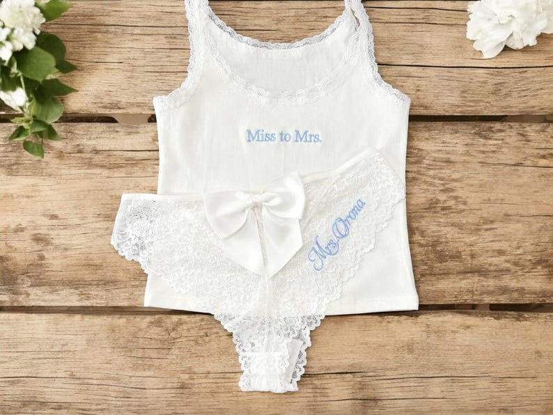 May include: White bridal lingerie set. A tank top with "Miss to Mrs." in light blue, and lace trimmed panties with a bow and "Mrs. Orona". The set is displayed on a wooden surface.