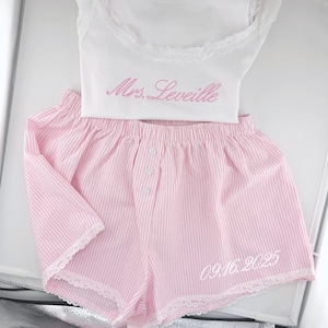 Puede incluir: Una camiseta sin mangas blanca con ribete de encaje y el texto bordado en rosa Mrs. Leveille, junto con pantalones cortos a rayas rosas y blancas con bragueta de tres botones, ribete de encaje blanco y la fecha bordada 09.16.2025.