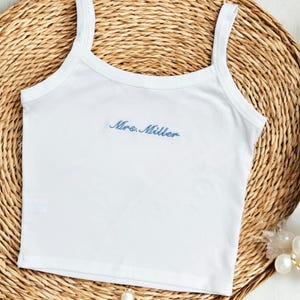 May include: A white sleeveless tank top featuring the text Mrs. Miller embroidered in blue script across the center of the chest. The garment has a scoop neckline and wide shoulder straps, presented in a clean, simple design for casual wear.