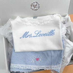 May include: A white shirt with a lace collar and the blue embroidered text Mrs. Leveille, paired with blue and white striped fabric featuring an embroidered monogram with a pink heart, all presented inside a white gift box with a thank you sticker.