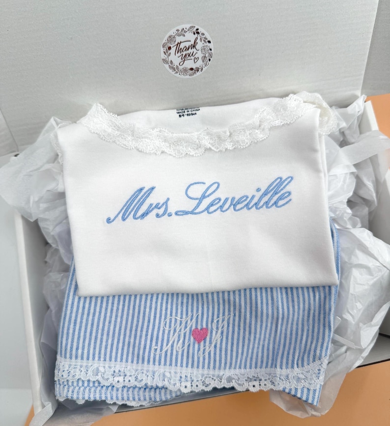 May include: A white shirt with a lace collar and the blue embroidered text Mrs. Leveille, paired with blue and white striped fabric featuring an embroidered monogram with a pink heart, all presented inside a white gift box with a thank you sticker.