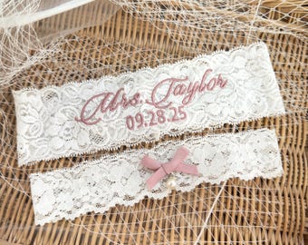 May include: Two white lace garters featuring floral patterns. The wider garter displays the embroidered text Mrs. Taylor and the date 09.28.25 in dusty rose thread. The narrower garter is accented with a dusty rose ribbon bow and a small pearl charm.
