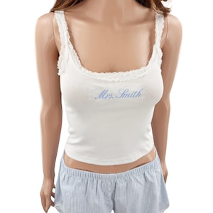 May include: A white tank top with lace trim along the scoop neckline and straps, featuring the text "Mrs. Smith" in light blue script. The tank top is paired with light blue and white pinstriped shorts with a button detail at the waist.