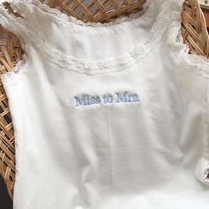May include: A white vest top with lace detailing around the neckline and straps. The words "Miss to Mrs." are embroidered in light blue. The top is made of a soft fabric.