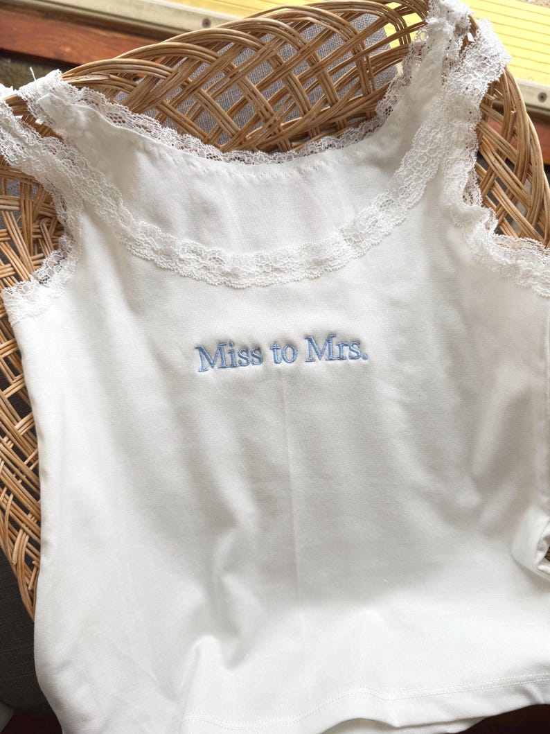May include: A white vest top with lace detailing around the neckline and straps. The words "Miss to Mrs." are embroidered in light blue. The top is made of a soft fabric.