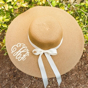 May include: A wide-brimmed sun hat in a natural tan color, featuring a circular crown and a broad brim. A white ribbon is tied around the crown, forming a bow with long flowing ends. The hat has a white monogram on the brim.
