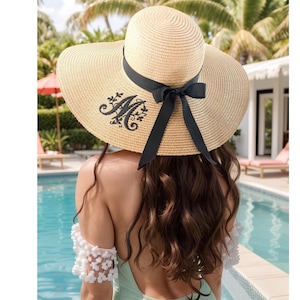 May include: A wide-brimmed straw sun hat with a black ribbon and bow. The hat features an embroidered black initial "M" with floral accents. The hat is worn by a person near a pool.