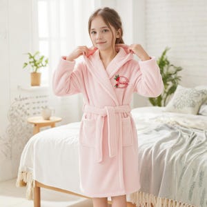 May include: A light pink hooded robe with a matching belt and pockets. The robe has the name "Daniela" embroidered on the chest, surrounded by a floral design. The robe is made of a soft, plush material.