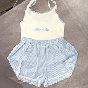 May include: A white tank top and blue striped shorts pajama set. The tank top has lace trim and the words "Miss to Mrs." in blue. The shorts have a button closure and lace trim. A small pink heart is visible on the shorts.