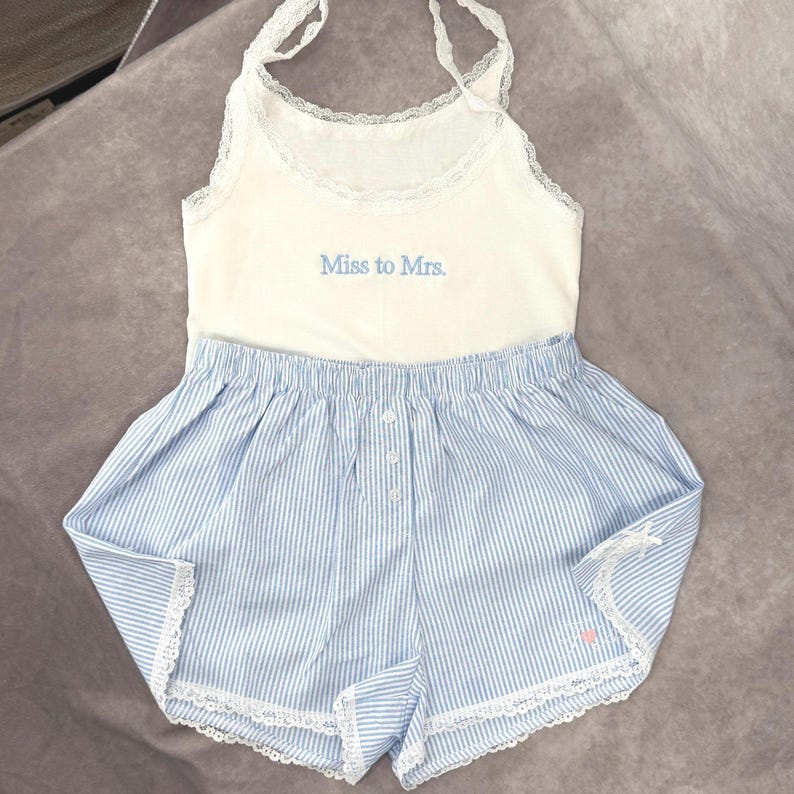 May include: A white tank top and blue striped shorts pajama set. The tank top has lace trim and the words "Miss to Mrs." in blue. The shorts have a button closure and lace trim. A small pink heart is visible on the shorts.